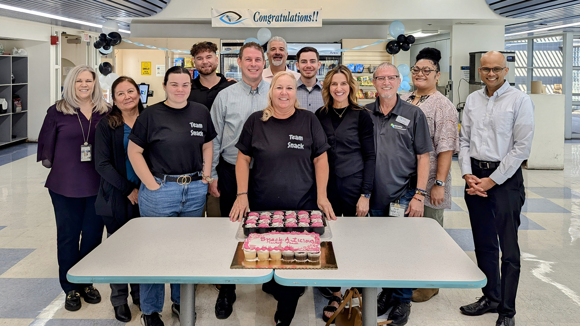 The BEN team Staff stand around trays of cupcakes smiling. Some are wearing shirts that say Team Snack. A banner with the BEN logo and congratulations hangs overhead.