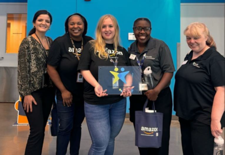 a diverse group of amazon management smile as they pose with vocational rehabilitation representatives and their plaque