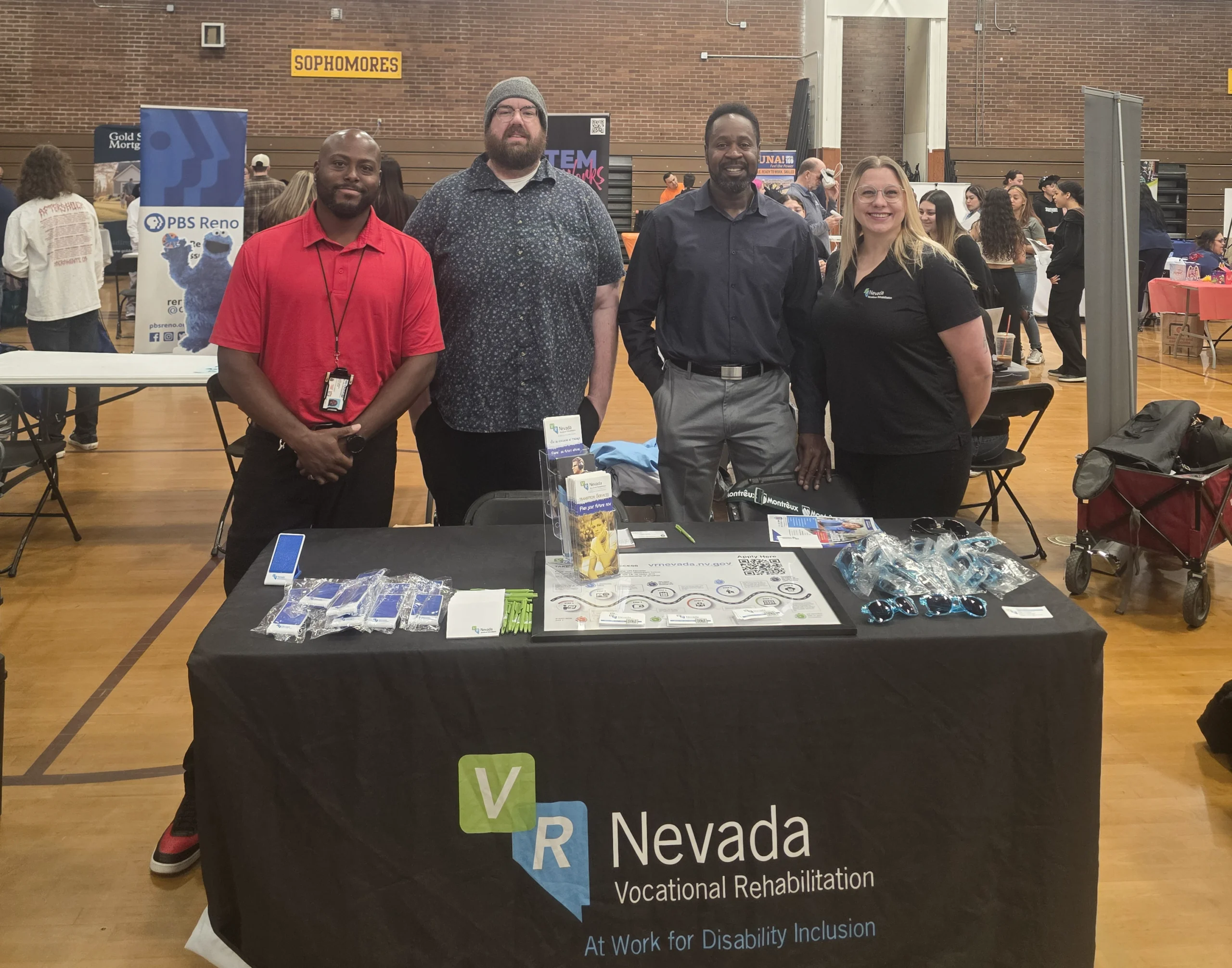 Four smiling VR Nevada representatives stand behind a booth at the Sparks High School info fair. The table features branded brochures, pens, and blue sunglasses under a "Vocational Rehabilitation" banner.