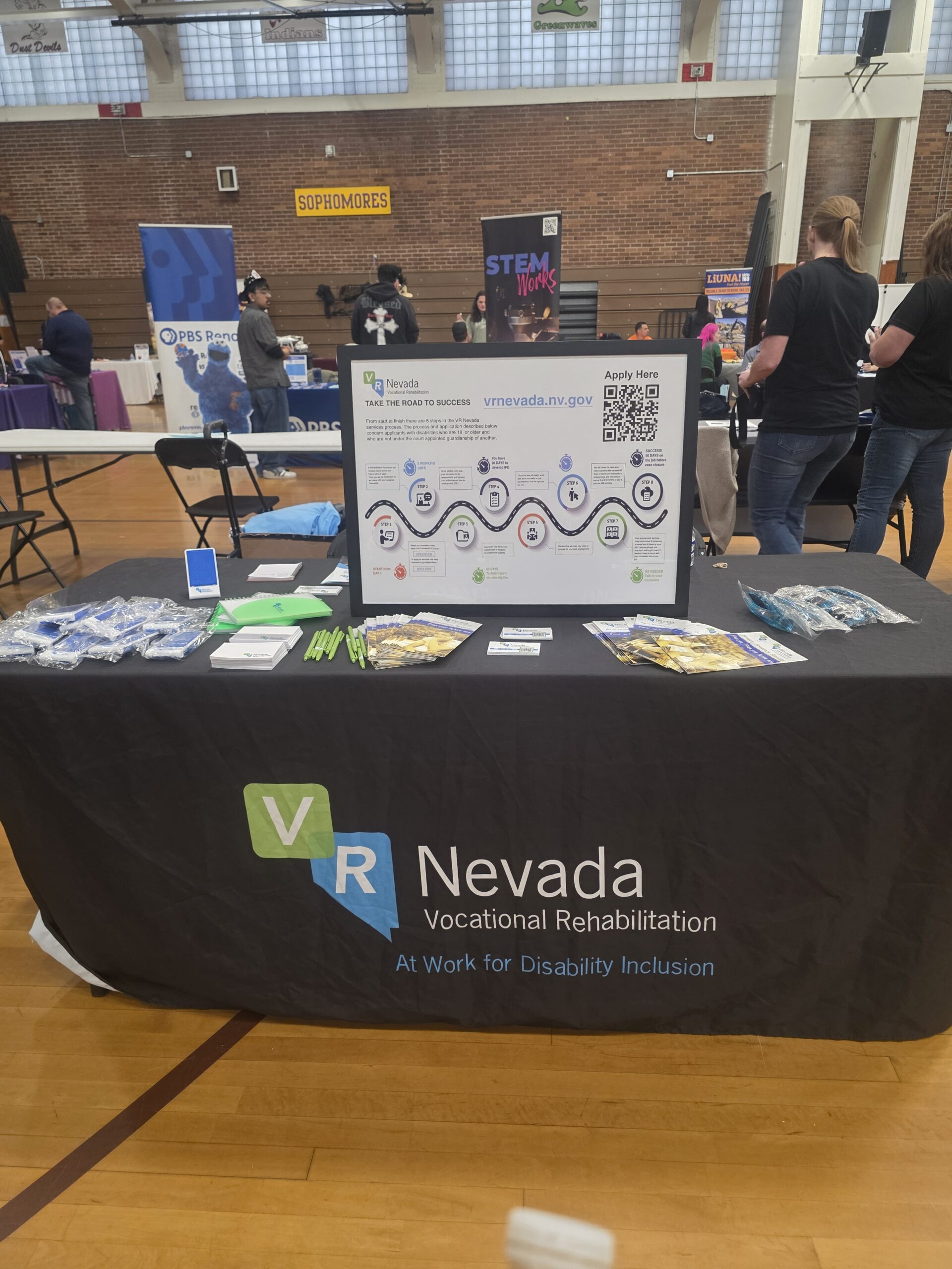 A close-up of the VR Nevada booth featuring a "Road to Success" infographic, a QR code for applications, and various promotional giveaways like green pens and brochures.