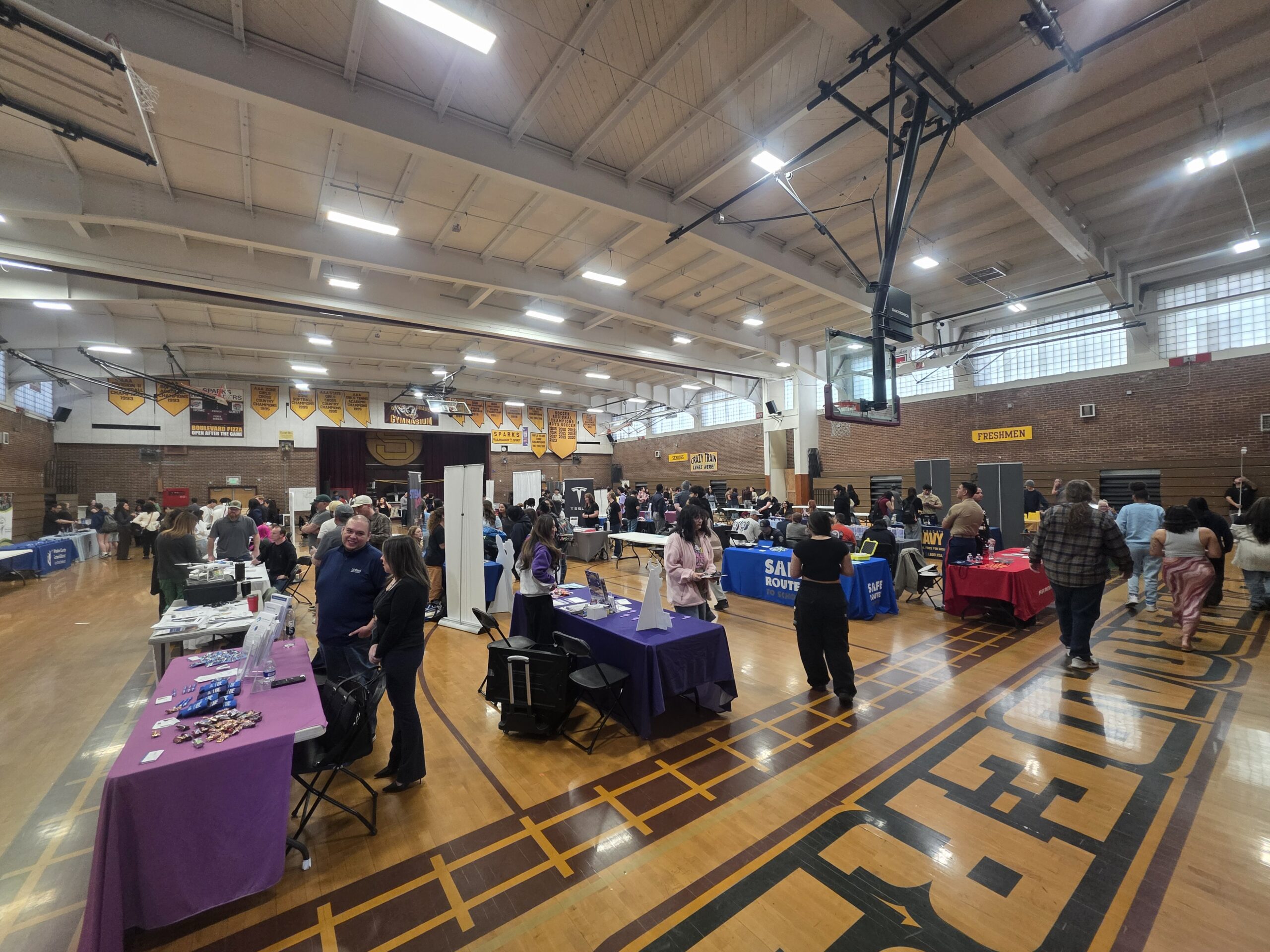 A wide view of a busy information fair inside the Sparks High School gymnasium. Students walk between rows of colorful booths set up on the wooden basketball court.