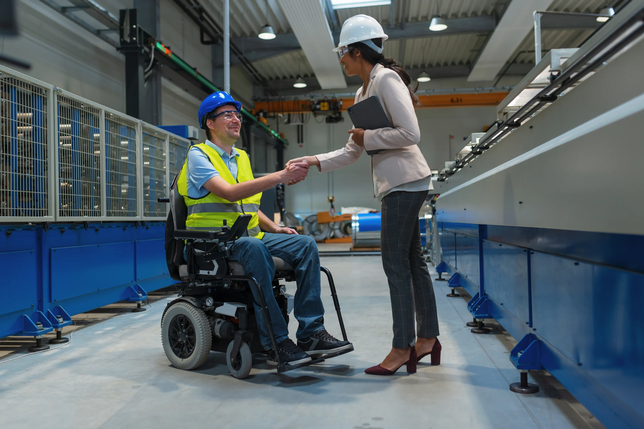 supervisor in white hard hat with clip board shakes hands with man in bkue hard hat and wheelchair