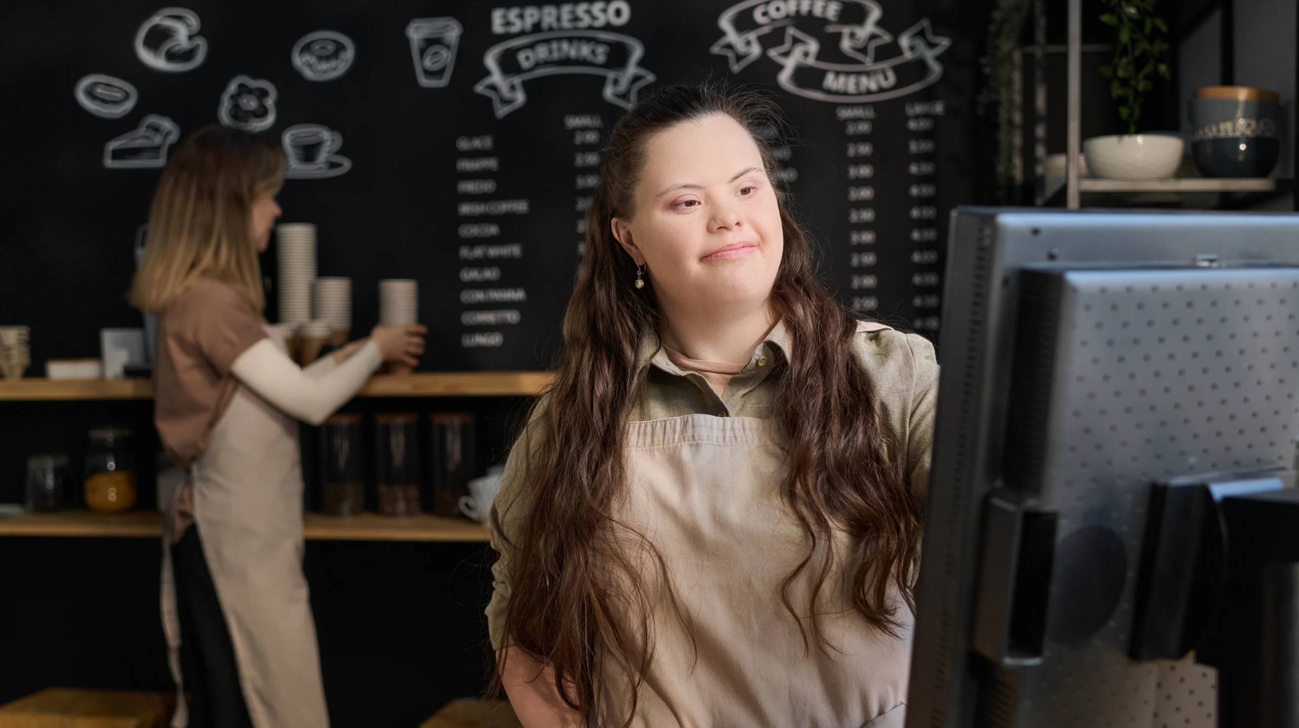Girl with Down syndrome looking at computer screen while standing by counter with young female colleague in workwear